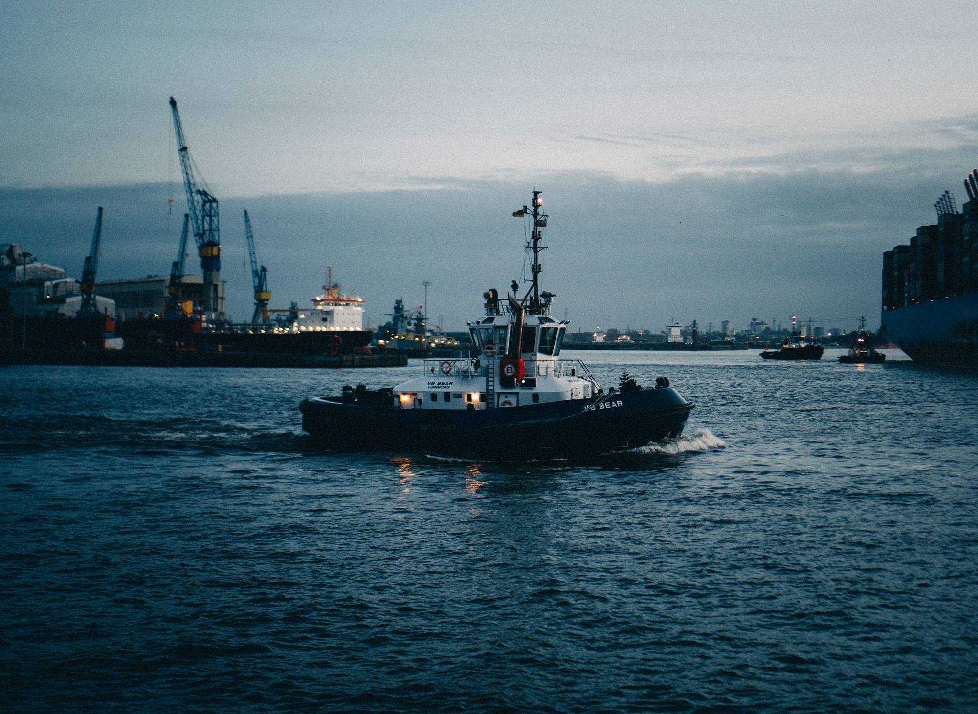 A tugboat crossing a busy port with cranes in the background