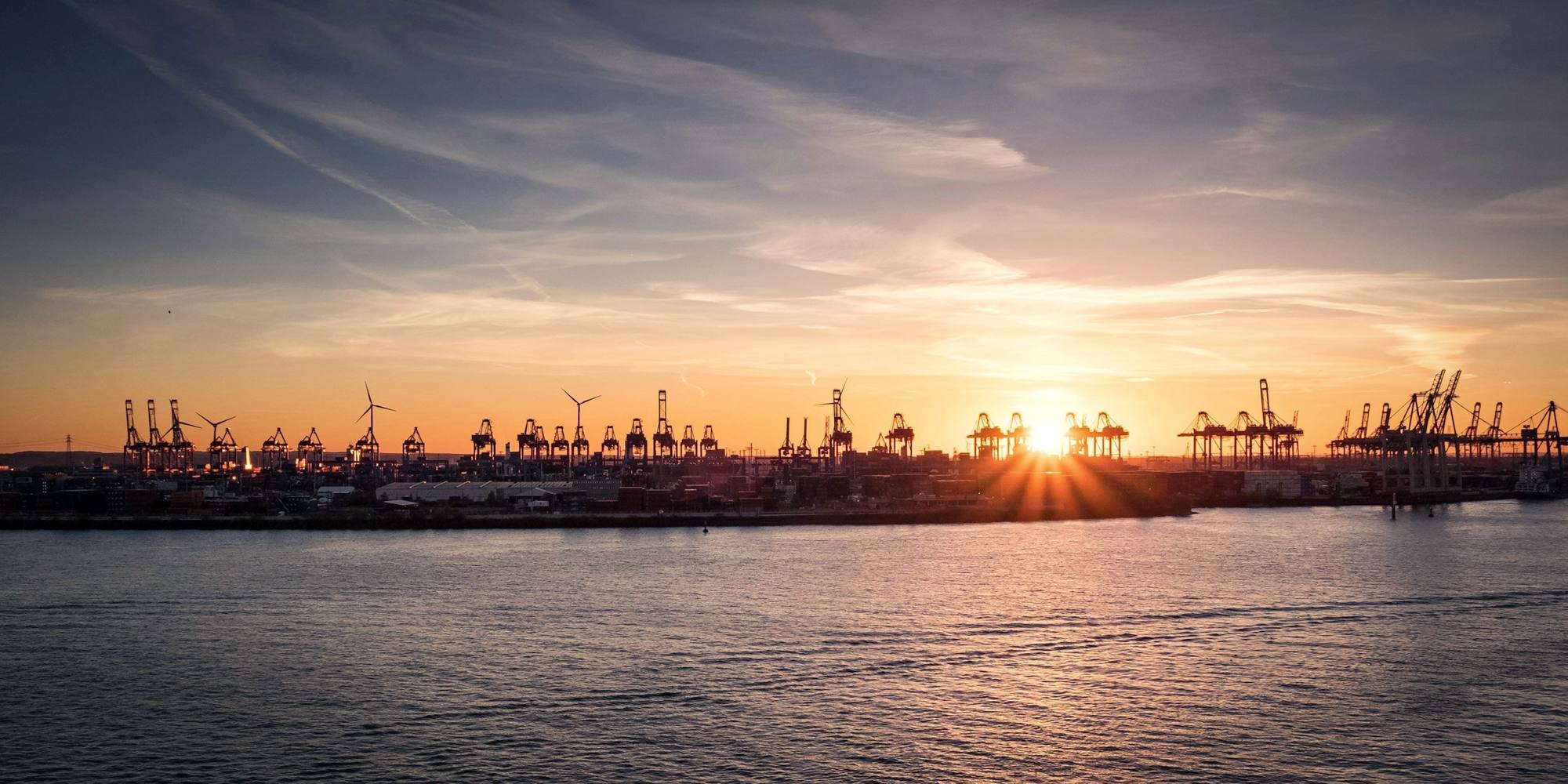 A container port at sunset, cranes silhouetted against the sky across calm water